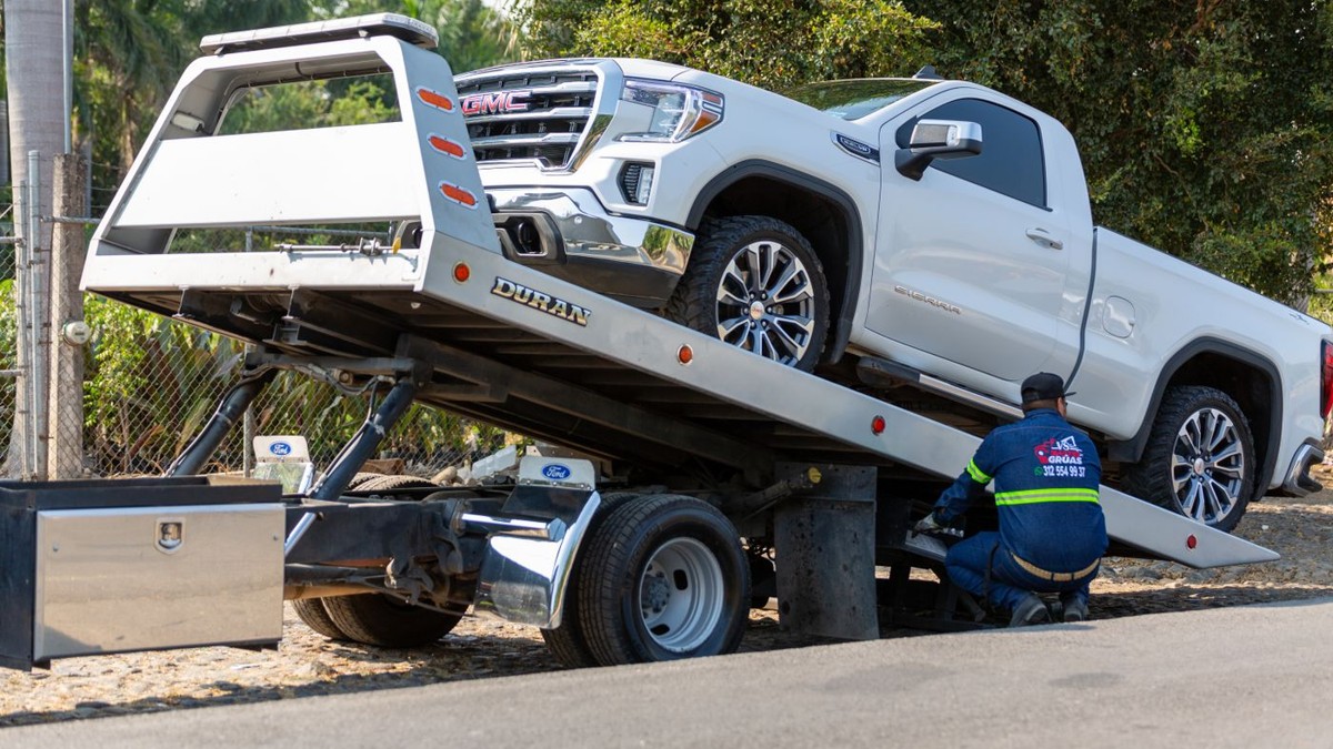 Stock photo: operator beside a tilted flatbed tow truck carrying a white GMC pickup.